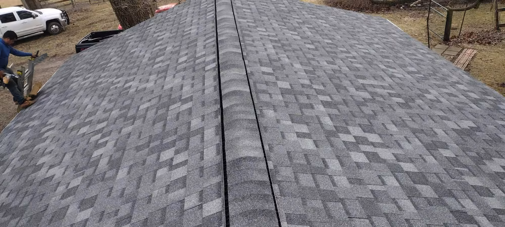 Long new gray architectural shingle roof with a ridge vent along the peak, a roofer and ladder at the left and a white truck below