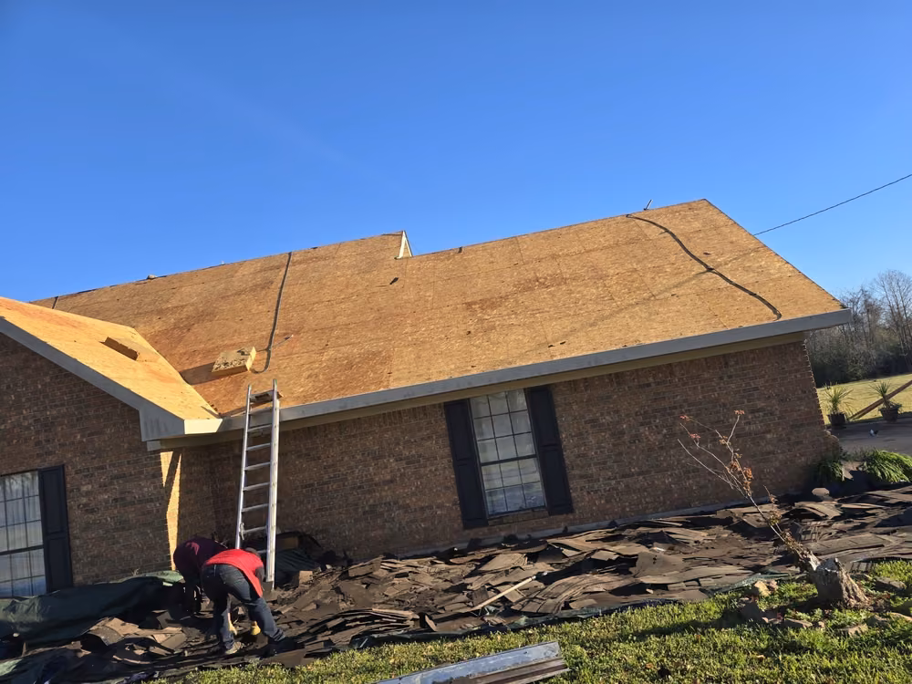 Brick house with the old roof torn off down to the decking while two workers clear old shingles off the ground tarps