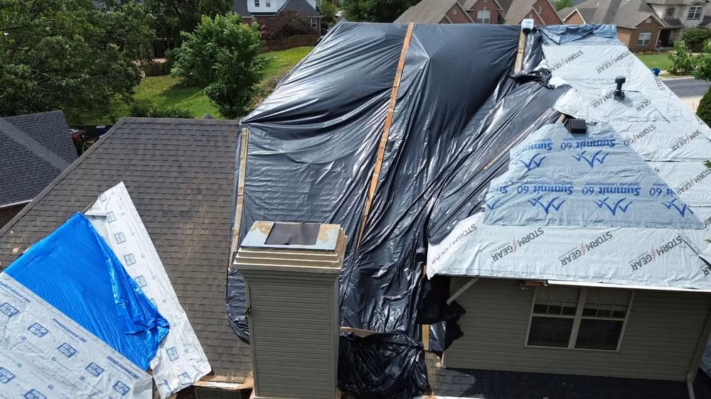 Storm-damaged roof covered with a black tarp and synthetic underlayment beside a chimney