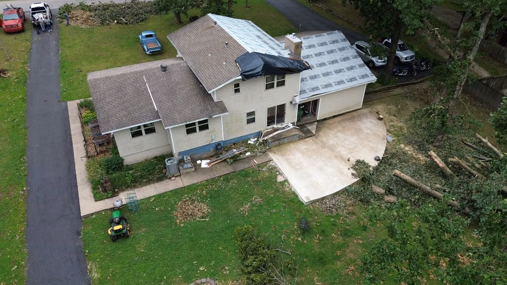 Drone view of a roof with a tarp over the section a fallen tree broke, the cut-up tree lying in the yard