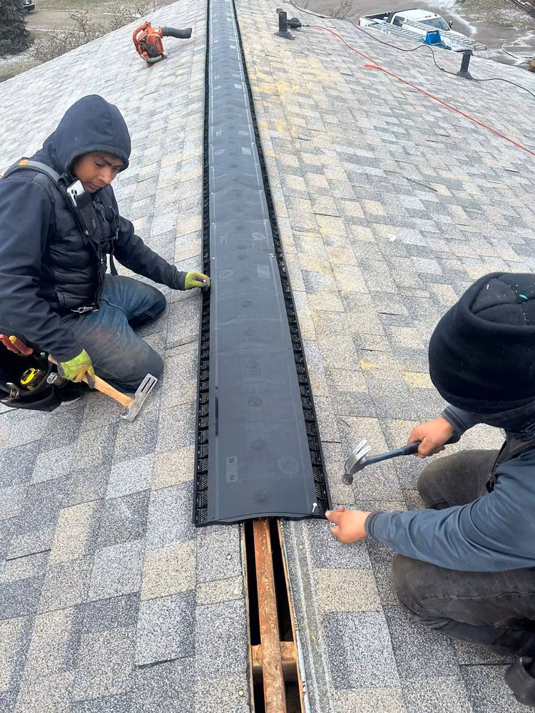 Two roofers nailing a new ridge vent along the peak of a shingle roof