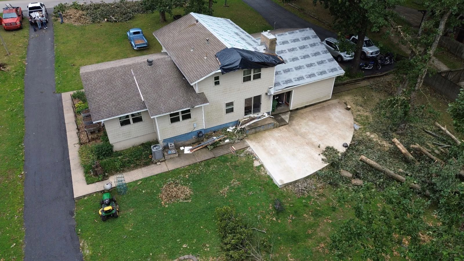 Aerial view of a storm-damaged house with a black tarp over the torn ridge and downed trees cut up in the yard